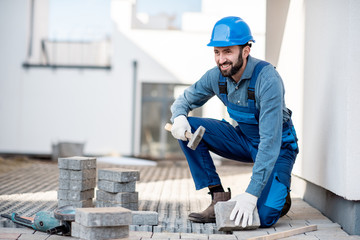 Builder in uniform laying paving tiles on the construction site with white houses on the background