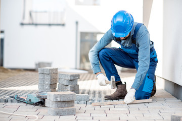 Builder in uniform laying paving tiles on the construction site with white houses on the background