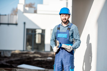 Porait of a builder in uniform holding hummer and paving tile on the construction site with white houses on the background