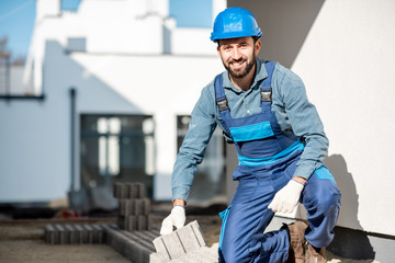 Portrait of a builder in uniform laying paving tiles on the construction site with white houses on...