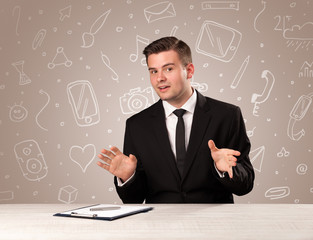 Young handsome businessman sitting at a desk with white mixed media icons behind him