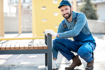 Handsome workman in uniform mounting a new bench on the playground outdoors
