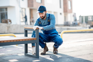 Handsome workman in uniform mounting a new bench on the playground outdoors