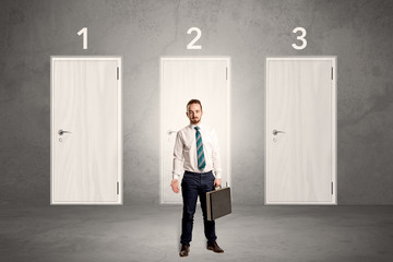 Obraz premium Businessman in grey room with three numbered white door behind his back 