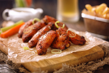 Roasted chicken wings served on a wooden board with glass of beer, french fries, carrot, celery and sause