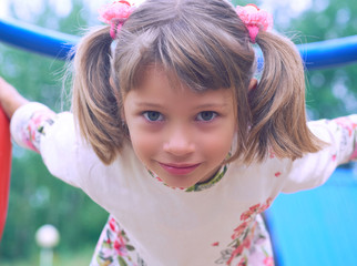 Portrait of cute Caucasian litte girl wearing white dress with flowers hanging on monkey bars on a summer day. Girl looking at camera smiling. Green leaves a seen in the background.