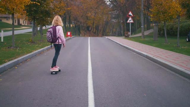Young pretty beautiful blond hipster woman in pink windstopper having fun riding skateboard longboard downhill on beautiful road in slow motion