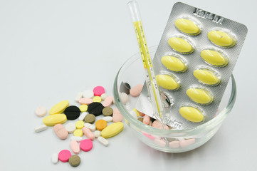 Assorted pharmaceutical medicine pills, tablets and capsules with thermometer on glass bowl