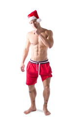 Athletic shirtless young man in Santa Claus hat standing to celebrate the Christmas season, isolated on white looking at camera