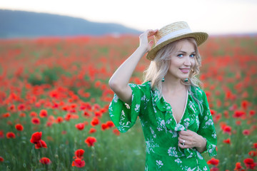 A handsome girl with long hair and natural skin, standing in a fiel of red poppies and holding a red poppy in hands, on nature landscape background. Horizontal view.