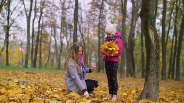Positive attractive mother helping her cute elementary age daughter arranging bouquet of yellow fallen maple leaves while relaxing together in autumn park. Joyful family with kid enjoying fall season.