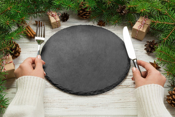 Perspective view girl holds fork and knife in hand and is ready to eat. Empty black slate plate on...