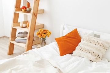 Home decor. Cozy fall bedroom interior: white wall, bed with white linen, light beige plaid, orange pillows, wooden rack, vase with yellow chrysanthemums flowers, pumpkins, candles. Autumn decoration.