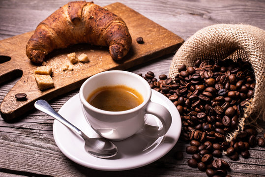 on the rustic wooden table, in the foreground, a cup of espresso coffee, a croissant with cereals, and roasted coffee beans.
