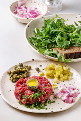 Beef tartare with quail egg in shell, cutting pickled cucumbers, capers, red onion, chives and arugula salad served in spotted ceramic plates with pepper mill on white marble table.