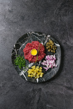 Beef tartare with quail egg in shell, cutting pickled cucumbers, capers, red onion, chives served in marble plates over black texture background. Flat lay, space