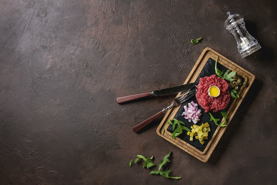 Beef tartare with quail egg in shell, cutting pickled cucumbers, capers, red onion, chives, arugula served on wooden black slate board with cutlery on dark texture background. Flat lay, space