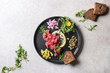Beef tartare with quail egg in shell, bread, cutting pickled cucumbers, capers, red onion, chives, arugula salad served in black ceramic plate over grey background. Flat lay, space