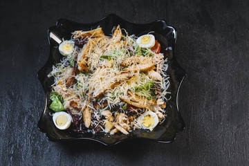 Delicious fresh caesar salad with chicken and eggs in a black plate on a black stone table background