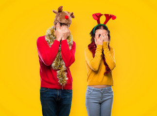 Couple dressed up for the christmas holidays covering eyes by hands and looking through the fingers on yellow background