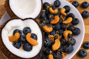 Healthy breakfast or snack in cut half coconut on wooden background. Yogurt in coconut bowl with blueberries, cashew nuts and granola.