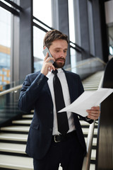 Young busy agent in formalwear reading paper and consulting client on smartphone by staircase