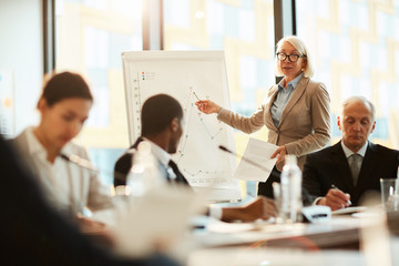 Mature blond female coach pointing at graph on whiteboard and explaining it to audience at seminar