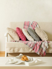 Beige sofa with plaid, colorful pillows (pink, grey, white) and coffee with croissantsin the living room.
