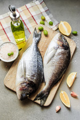Fresh raw dorado or sea bream fish on a wooden kitchen board with lemon slices, spices, Himalayan pink salt, olive oil. Gray background. Mediterranean Kitchen.