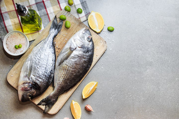 Fresh raw dorado or sea bream fish on a wooden kitchen board with lemon slices, spices, Himalayan pink salt, olive oil. Gray background. Mediterranean Kitchen. Top view. Place for text.