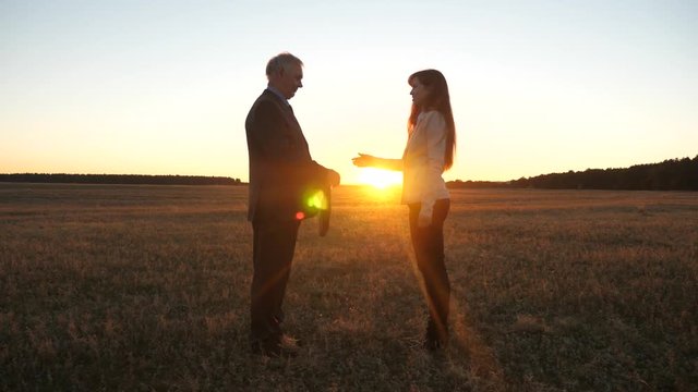 Business lady transmits briefcase with documents to an elderly business man and makes deal with him with handshake and smile against sunset in sun.