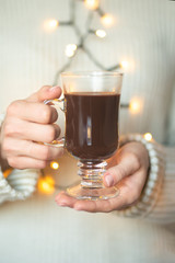 A woman in a bright warm woolen holds hot chocolate in her hands, and a burning Gerland against the background, copy space, selective focus. Christmas, new year holiday concept