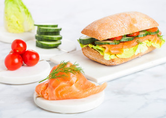 Fresh healthy salmon sandwich with lettuce and cucumber and tomatoes on marble board on white marble background. Breakfast snack.
