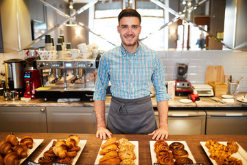 Young bakery clerk in apron standing by counter with large assortment of fresh pastry