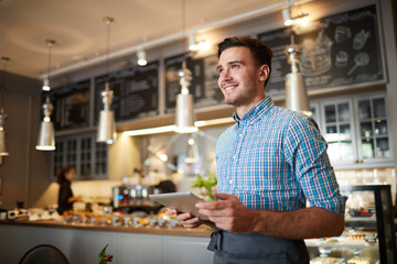 Young cheerful man in workwear standing in modern cafeteria with counter on background