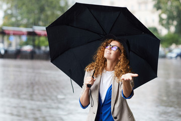 Happy girl looking upwards from under her black umbrella while enjoying rain during walk - obrazy, fototapety, plakaty