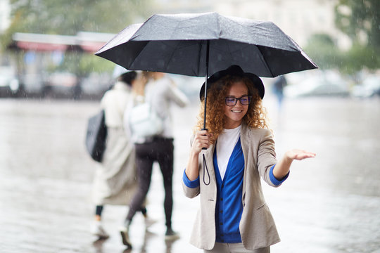 Young charming woman in stylish casualwear walking under umbrella and playing with raindrops
