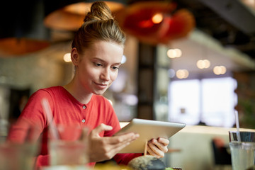 Contemporary young female blogger with tablet siting in cafe and recording video for her internet channel subscribers