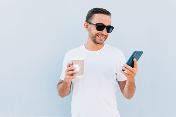 Happy handsome guy in sunglasses is looking at screen of mobile phone isolated on blue background