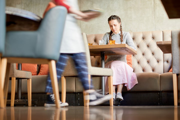 Pretty girl sitting on sofa in cafe and scrolling in smartphone while waiting for her order