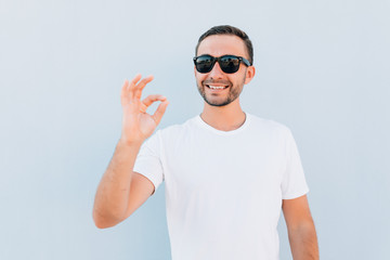 Naklejka premium Young smiling man in sunglasses having happy look, gesturing, showing OK sign or showing okay gesture with his fingers on blue background