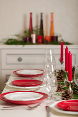 Merry Christmas and Happy New Year! Тable setting festive decor - white and red dishes, red candles in the candlesticks, juniper branches and fir cones.