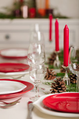 Merry Christmas and Happy New Year! Тable setting festive decor - white and red dishes, red candles in the candlesticks, juniper branches and fir cones.