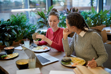 Two casual girls sitting by table in front of laptop, having fast food and discussing home assignment after lessons