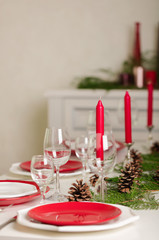 Merry Christmas and Happy New Year! Тable setting festive decor - white and red dishes, red candles in the candlesticks, juniper branches and fir cones.