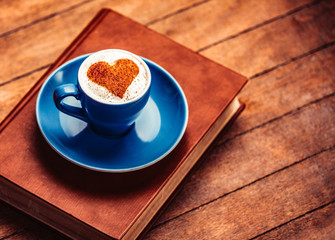 Blue cup of coffee with heart shape symbol and book on wooden table and background