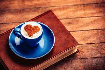 Blue cup of coffee with heart shape symbol and book on wooden table and background