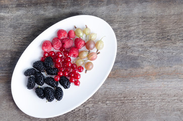 Fresh berries on white plate. Mulberries, currants, gooseberries, raspberries. Wooden background with copy space