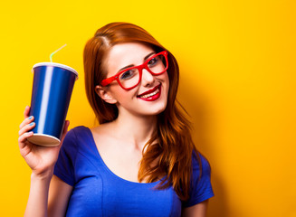 Style redhead girl in blue dress and glasses with drink on yellow background.
