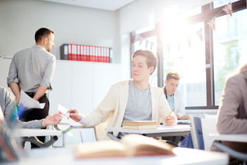 Casual guy passing over note with crib to his groupmate behind teacher back